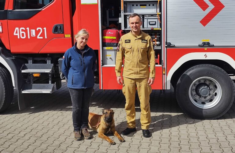 Zdjęcie Pani Prezes z Komendantem i psem ratowniczym na tle samochodu bojowego