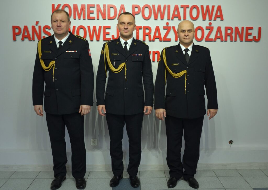 Trzech odznaczonych i wyróżnionych strażaków stojących na tle napisu komendy powiatowej Straży Pożarnej w Łęcznej